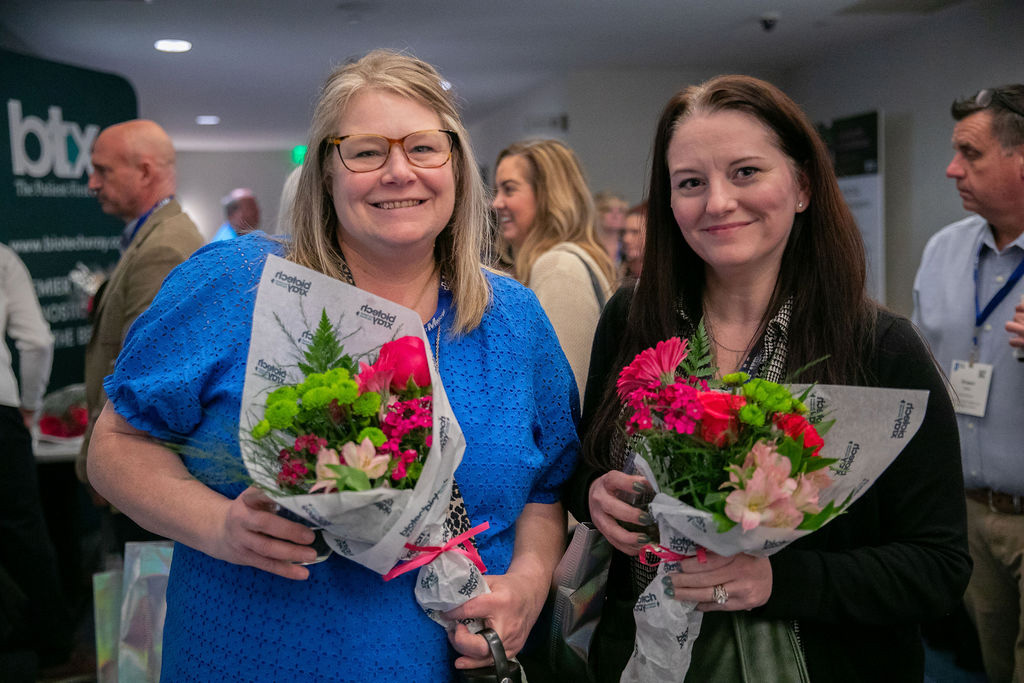 two woman smiling at the camera while holding flower bouquets